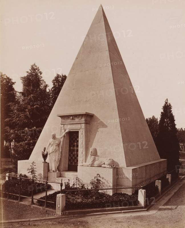 Crematorium, Cimitero Monumentale, Milan, Italy, photograph, Classical marble building, the Crematorium of the Cimetero Monumentale cemetery, Italy., caption: '9649. MILANO. Cimitero Monumentale, Crematoio, architetto Maciacchini.', caption: '(EDIZIONI BROGI)', photographer: Edizioni Brogi, Florence, publisher: Giacomo Brogi, c. 1865 - c. 1890, Tuscany, pasteboard, paper, albumen print, height 249 mm x width 200 mm, height 492 mm x width 330 mm, Lombardy, A large stone pyramid structure fills most of the image, with smooth, angled faces and a sharp apex. At the base of the pyramid is a decorated doorway framed by carved relief panels and a horizontal lintel. A life-sized draped female statue stands beside the doorway, one arm raised to touch the frame and the other resting near a tall vase on a pedestal. In front of the doorway, a recumbent sphinx sculpture lies on a raised platform, facing outward. The pyramid rests on a stepped base surrounded by low stone walls and a wrought metal f...
