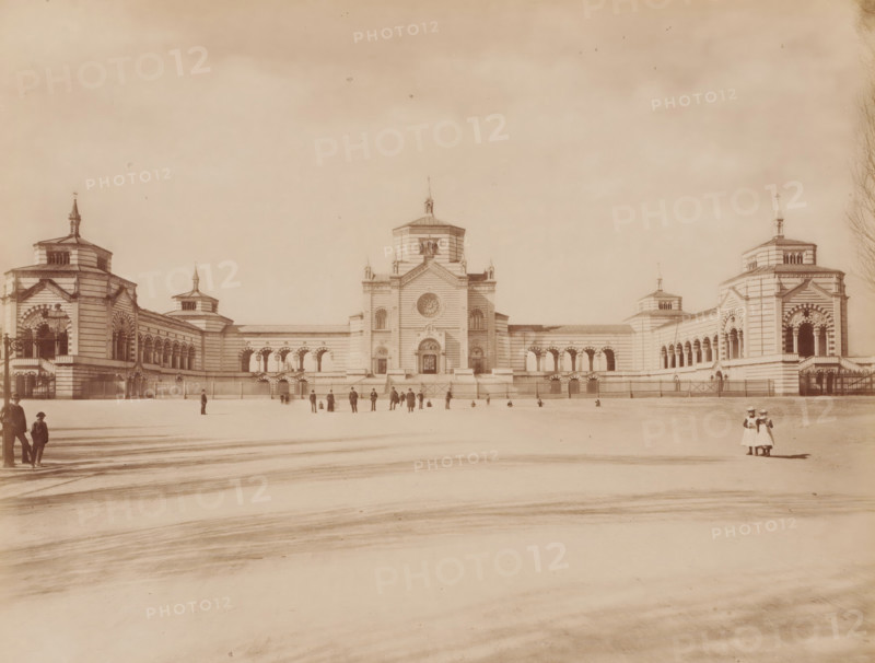 Veduta generale del. famedio, Cimitero Monumentale, Milan, Italy, photograph, Italy., caption: '9646. MILANO - (Cimitero Monumentale) Vedura generale del. Famedio', caption: '(EDIZIONI BROGI)', photographer: Edizioni Brogi, Florence, publisher: Giacomo Brogi, c. 1860 - c. 1890, Tuscany, pasteboard, paper, albumen print, height 252 mm x width 198 mm, height 330 mm x width 510 mm, Lombardy, A wide pale photograph of a large symmetrical stone building with a central tower flanked by lower wings and matching end pavilions, each pavilion topped with a small spire; the façade shows arched openings, striped stonework, decorative columns, and a large circular window in the center tower above an ornate main entrance; a low fence runs along the front of the building with several pedestrian gates; an open paved foreground stretches to the camera with faint surface markings and long shadows; small groups of people and a few solitary figures are scattered near the building and along the foreground,...