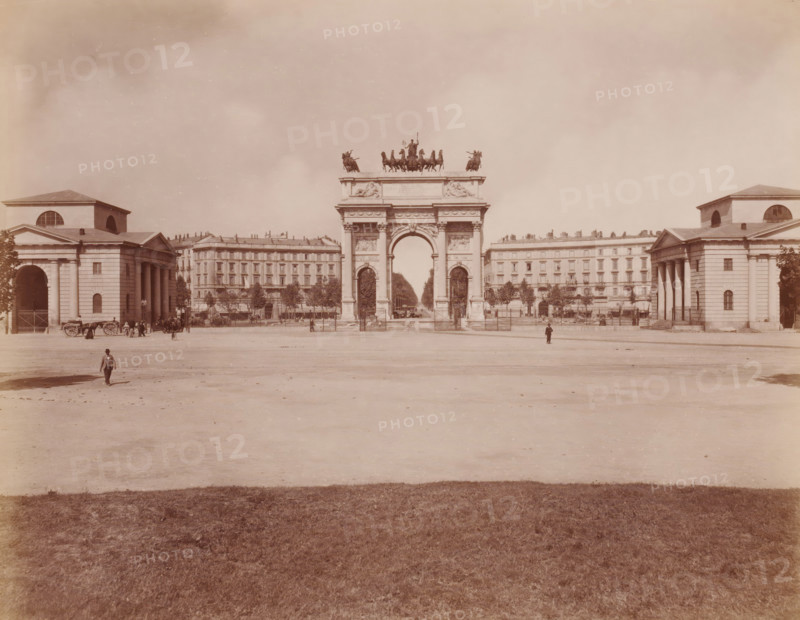 Veduta d'assieme con Arco della Pace, Milan, Italy, photograph, Italy., caption: '12291. MILANO - Veduta d'assieme con l 'Arco della Pace.', caption: '(EDIZIONI BROGI)', photographer: Edizioni Brogi, Florence, publisher: Giacomo Brogi, c. 1860 - c. 1890, Edizioni Brogi, Giacomo Brogi, photographer, publisher, Tuscany, pasteboard, paper, albumen print, height 250 mm, width 200 mm x width 510 mm, height 330 mm, Lombardy, A wide open plaza with a central monumental arch topped by a sculptural group of horses and figures, flanked on either side by classical buildings with columns and arched openings, a line of multi-story buildings behind the arch, trees and fencing near the buildings, a few people and horse-drawn carts near the side buildings and a couple of solitary figures in the open area, and a foreground of grass leading to the bare expanse of the plaza.