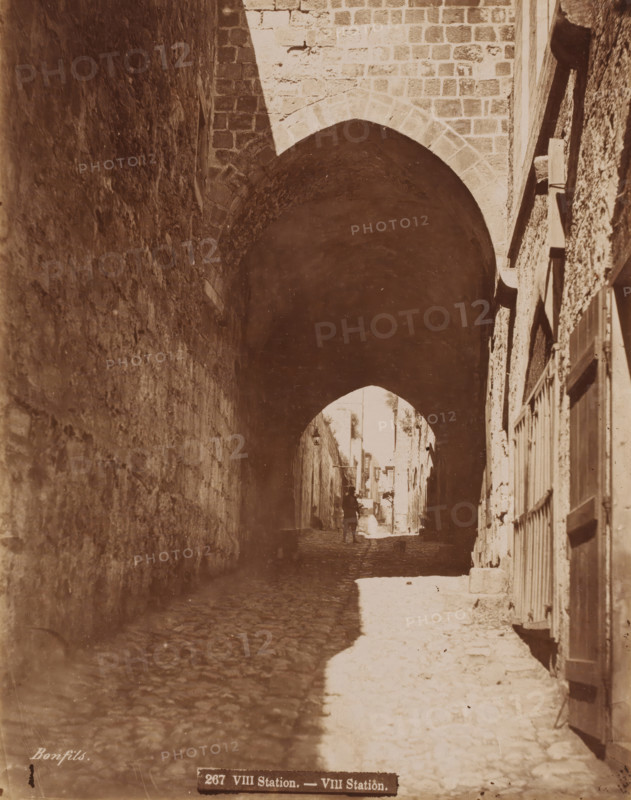 VIII Station', eighth station in the Passion of Christ?, Jerusalem?, Palestine?, photograph, View of a gate, signature: 'Bonfils', inscription: '267 VIII Station. - VIII Station.', photographer: Maison Bonfils, c. 1880 - c. 1900, Maison Bonfils, photographer, pasteboard, paper, albumen print, height 277 mm, width 218 mm x width 344 mm, height 394 mm, A narrow cobblestone street runs beneath a large stone archway, with tall textured stone walls on either side; light falls on part of the right side of the street creating a bright patch on the ground while the left side remains in shadow, and a small silhouetted figure is visible in the distance under the arch with a few indistinct details of buildings and doorways beyond.