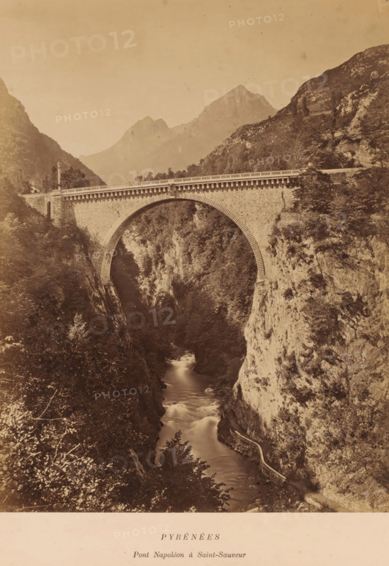 PYRÉNÉES, Pont Napoléon à Saint-Sauveur', Luz-Saint-Sauveur, France, photograph, Frontal view of the Pont Napoleon, an arch bridge built in 1860., caption: 'PYRÉNÉES, date: '11, 12 October 1884.', caption: 'LAFONT LIBRAIRE ÉDITEUR, 40 ALLÉE D'ETIGNY A LUCHON', vendor: Lafont, Luchon, 1884-10-11 - 1884-10-12, Lafont, photographer, vendor, pasteboard, paper, albumen print, albumendruk, height 276 mm, width 212mm x width 338mm, height 398 mm, Pyrenees, A stone arch bridge spans between two steep rocky cliffs above a narrow river flowing through a deep gorge, the bridge showing textured masonry and a balustrade along its roadway, with vegetation and trees clinging to the cliff faces and surrounding the gorge, a winding path or retaining wall visible along the right cliff beside the water, distant mountain peaks rising in the background under a pale sky.