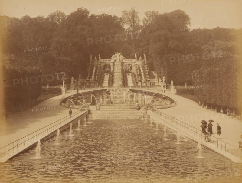 Parc de St. Cloud, La Grande Cascade', Paris, photograph, View of a long symmetrical pond with fountains and some people with umbrellas, parasols?, France, check stamp: 'Photographic ? Company, J.KUHN, PARIS, 220 170174 220, RUE DE RIVOLI', inscription: 'No. 247 PARC de St. Cloud La Grande Cascade X. Phot.', publisher: J. Kühn (publisher), c. 1880 - c. 1900, J. Kühn (publisher), publisher, pasteboard, paper, albumen print, height 222 mm, width 279 mm x width 401 mm, height 304 mm, A wide formal water feature stretches toward a large tiered stone cascade and sculptural terrace set against a densely treed hillside, with parterres and hedges flanking the central axis; stone balustrades line the long reflecting basin, small jets of water arcing into the pool along the sides, and stepped waterfalls descend from the ornate terrace into lower basins; classical statues stand on plinths at the terrace and along the walkways; people in period dress walk or stand on the paths beside the water, so...