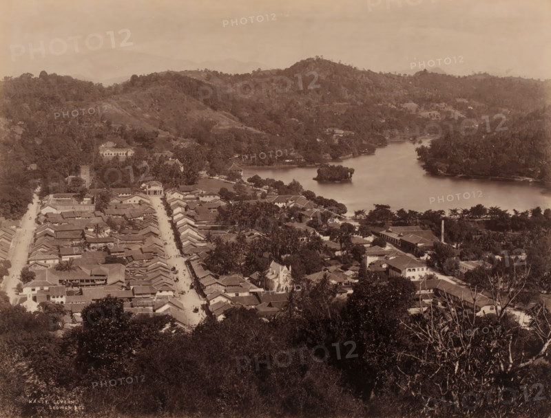 View of Kandy, Sri Lanka, photograph, View of the city of Kandy in the district of the same name, Sri Lanka., inscription: 'Kandy, Ceylon., Scowen & Co.', Ceylon', photographer: Charles T. Scowen & Co., Kandy, c. 1870 - c. 1890, Charles T. Scowen & Co., photographer, South Asia, pasteboard, paper, albumen print, height 213 mm, width 281 mm x width 407 mm, height 323 mm, A sepia aerial view showing a town with rows of tiled roofs and narrow streets leading toward a waterfront, a river or lake curving through the middle ground with a small vegetated island near its center, tree-lined banks and scattered buildings along the shoreline, clusters of trees and palms interspersed among houses, a prominent church-like building with a pointed roof near the street, larger structures set back amid greenery, a wooded foreground with dense foliage, and rolling forested hills rising in the background under a hazy sky.