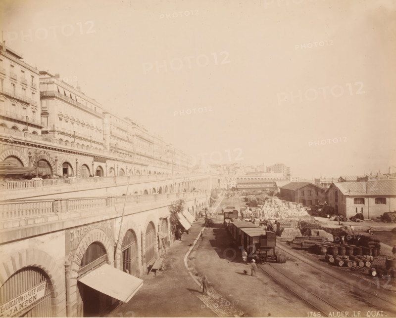 Current Boulevard Ernesto Che Guevara, Algiers, Algeria, photograph, View of the boulevard and the freight transshipment below., inscription: '1748. ALGER LE QUAI.', c. 1866 - c. 1890, photographer, paper, pasteboard, albumen print, height 213 mm, width 265 mm x width 440 mm, height 324 mm, North Africa, A wide riverside scene showing a long row of multistory buildings with arched lower levels and balconies along a raised promenade, a roadway and rail tracks running beside them, freight cars on the tracks, stacks of large sacks and barrels piled near warehouse-like sheds with pitched roofs, scattered carts and crates, several small groups of people and blurred figures moving along the road, lamp posts and railings along the elevated walkway, and distant low buildings and arches fading into the background.