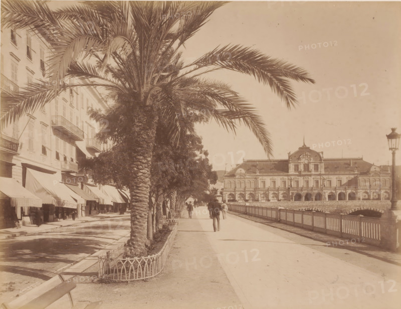 Boulevard, France ?, photograph, View of the boulevard with shops on the left, a bridge and behind it a large building on the right, with shop fronts displaying 'Renaissance' =museum?, and palm, other trees., c. 1880 - c. 1910, photographer, paper, pasteboard, albumen print, height 208 mm, width 270 mm x width 447 mm, height 326 mm, A sepia-toned street scene with tall palm trees lining a wide promenade alongside a railing and water, an ornate multi-story building with arched openings and decorative roofline visible across the promenade, a row of buildings with awnings and balconies on the left, a few pedestrians walking along the pathway, a lamppost near the right edge, and shadows cast on the ground.