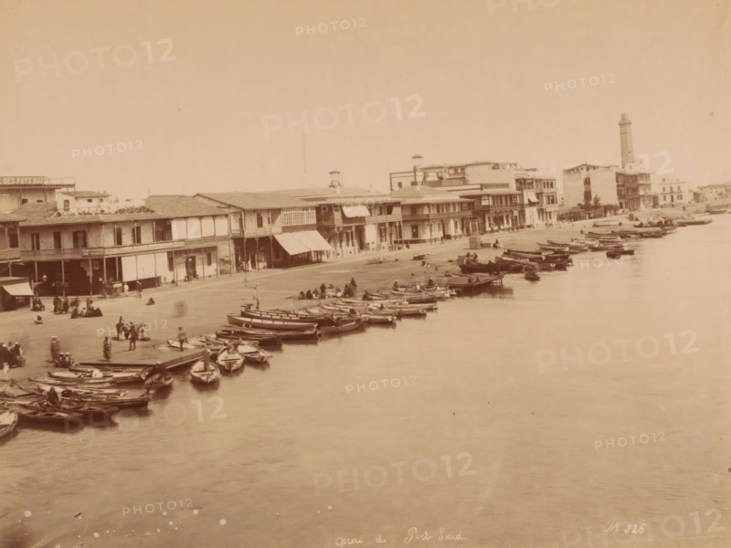 Quai de Port Said, Egypt, photograph, Egypt., inscription: 'quai de Port Said No. 326', photographer: attributed to Hippolyte Arnoux, c. 1860 - c. 1891, Attributed to, pasteboard, paper, albumen print, height 210 mm, width 278 mm x width 459 mm, height 323 mm, North Africa, Port Said, A waterfront scene with a row of two-story buildings along a shoreline, awnings and covered balconies on some facades, a lighthouse tower visible toward the right, numerous small wooden boats moored along the water's edge and floating on calm water, groups of people standing and sitting on the shore and near the boats, a wide expanse of water in the foreground and an open sky above.