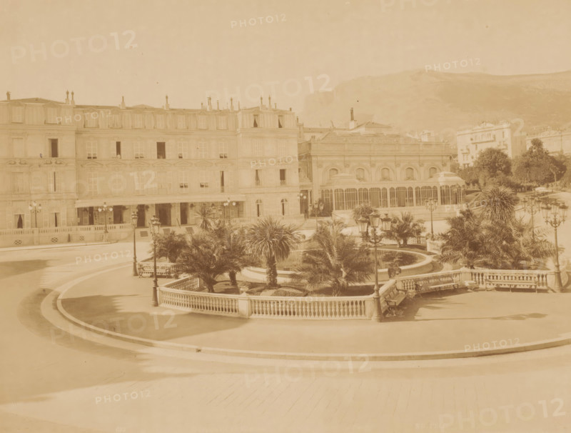 Square with roundabout with palm trees and pond in the middle and houses around it, photograph, Square with roundabout with palm trees and pond in the middle and houses around it., inscription: '622 ...', inscription: 'Phot. ND', signature: 'ND Phot.', photographer: Étienne Neurdein, c. 1880 - c. 1900, pasteboard, paper, albumen print, height 210 mm, width 276 mm x width 474 mm, height 323 mm, A wide view of an ornate building facade with multiple rows of windows and balconies adjacent to a structure with large arched windows, set behind a curved stone balustrade enclosing a landscaped area with palm trees, trimmed shrubs, and a circular water feature. Decorative street lamps with multiple globes line the paved terraces and paths, and benches sit along the balustrade. In the foreground a broad paved promenade sweeps in curved bands, and in the background low hills rise behind the buildings. The image has a uniform sepia tone.