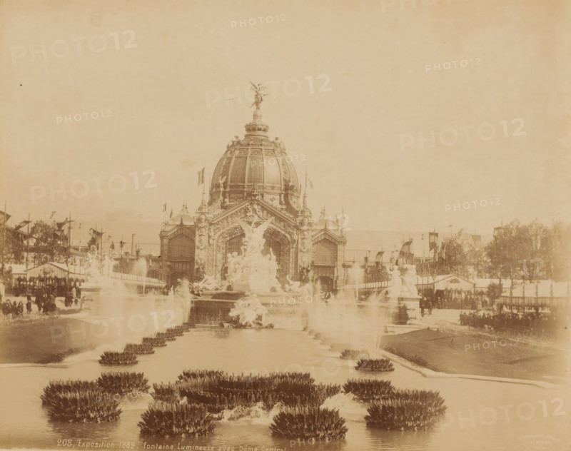 World Exhibition 1889 ('Exposition 1889 - fontaine Lumineuse avec Dôme Central.'), Paris, photograph, France., check stamp: 'Photographic Company, J. Kühn, 220 170174 Rue de Rivoli', inscription: '208, Exposition 1889 - fontaine Lumineuse avec Dôme Central', publisher: J. Kühn (publisher), 1889, photographer, pasteboard, paper, albumen print, albumendruk, height 215 mm, width 275 mm x width 446mm, height 343mm, World's Fair 1889 (Paris), A sepia-toned photograph shows a grand domed building with ornate architectural details and sculptural decorations; a winged statue tops the dome and additional statues and carvings adorn the façade. In front of the building a large decorative fountain complex features multiple sculpted figures and flowing water, with jets and cascades visible. A long reflecting pool or canal stretches toward the foreground, lined with low plant clusters emerging from the water. Flanking the central scene are procession-like rows of flags and banners, temporary structu...