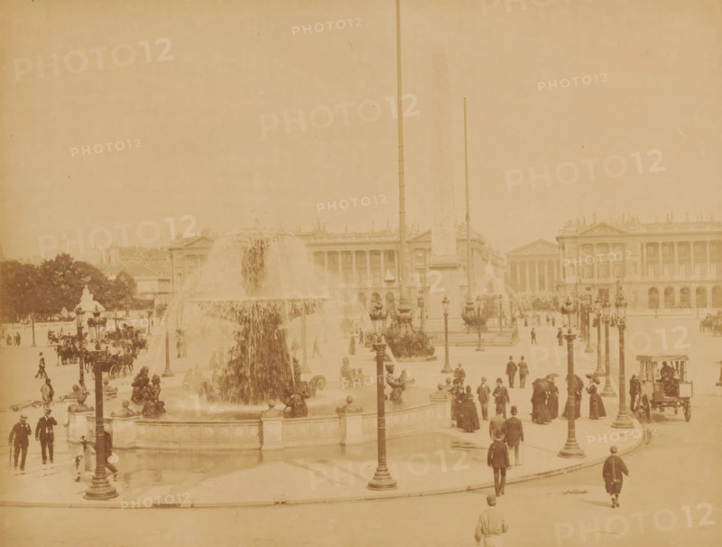 Place de la Concorde, Paris, photograph, View of the square with a large fountain in the middle and passers-by., inscription: '79 PARIS - Place de la Concorde', number: '19.', signature: 'X. Phot.', c. 1870 - c. 1900, photographer, pasteboard, paper, albumen print, albumendruk, height 211 mm, width 273 mm x width 446 mm, height 343 mm, A wide plaza scene shows a large ornate fountain with water cascading from its upper basin into a lower pool, surrounded by sculpted figures and decorative railings, with several tall lampposts positioned around the fountain and plaza; an obelisk rises behind the fountain flanked by flagpoles, and classical buildings with columns and detailed facades form the background; many people in period clothing are walking, standing, and sitting throughout the plaza, with horse-drawn carriages and a few clustered trees visible at the edges.