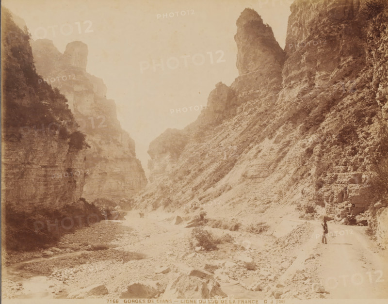 Ligne du Sud de la France, Gorges de Cians', France, photograph, inscription: '7166. GORGES DE CIANS - LIGNE DU SUD DE LA FRANCE', inscription: 'GJ photo.', photographer: Jean Gilletta, c. 1880 - c. 1900, Jean Gilletta, photographer, paper, pasteboard, albumen print, height 217 mm, width 275 mm x width 317 mm, height 387 mm, A sepia-toned scene of a narrow rocky canyon with steep cliff faces on both sides, showing layered rock textures and sparse vegetation clinging to the slopes, a dry rocky riverbed running through the canyon floor with scattered boulders and small pools of water, a winding dirt path along one side of the canyon, and a solitary person standing on the path carrying an object over their shoulder.