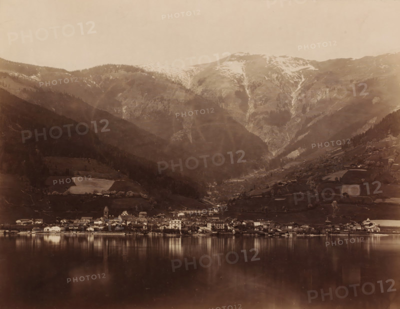 Landscape with a lake and mountains and a place on the shore of the lake, photograph, c. 1880 - c. 1900, photographer, pasteboard, paper, albumen print, height 205 mm, width 265 mm x width 373 mm, height 300 mm, A lakeside town with clustered buildings along the shore reflected in calm water, set against steep, forested hills and high mountains with patches of snow and visible gullies, terraces and scattered farm plots on the slopes, and a sky above.