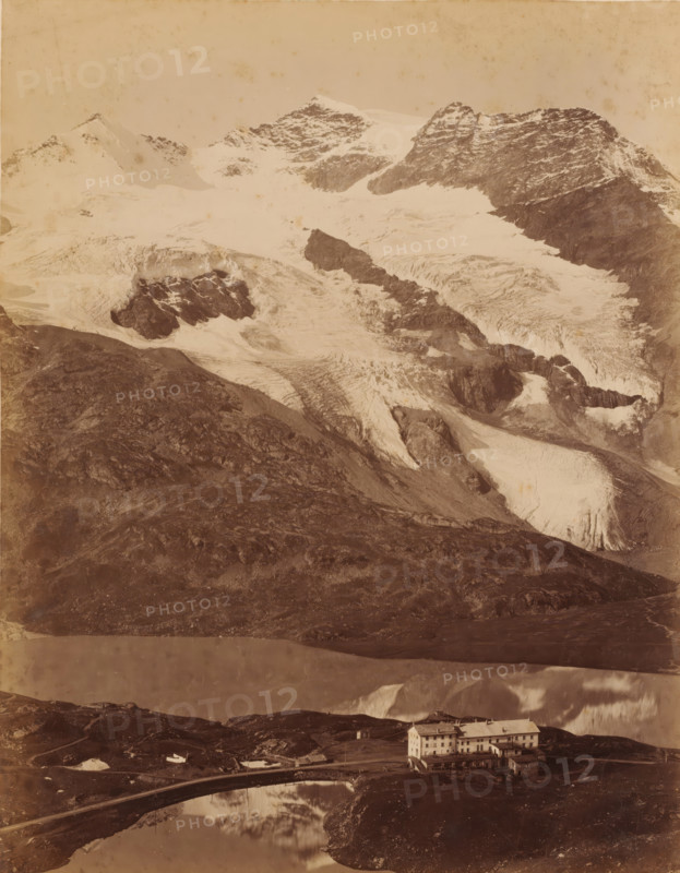 Bernina Pass, Switzerland, photograph, The Bernina Pass connects the Veltlin with the Engadin., inscription: 'Cauma See (Flims)', number: '21', c. 1890 - c. 1910, photographer, pasteboard, paper, albumen print, albumendruk, height 266 mm, width 205 mm x width 300 mm, height 379 mm, A wide mountain scene shows a steep rocky foreground leading up to snow-covered ridges and glaciers with visible crevasses and dark exposed rock bands, a pale sky above, a reflective body of water in the lower middle reflecting parts of the landscape, a curved narrow bridge crossing the water, and a multi-story pale building with a pitched roof and several small outbuildings situated near the water's edge.