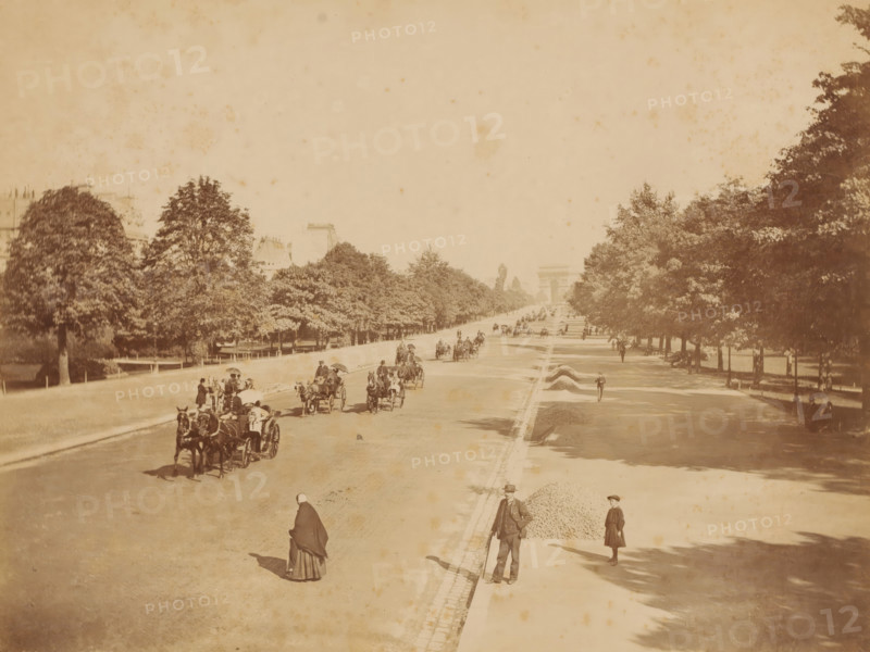 Perspective de l'Avenue du Bois de Boulogne', Paris, photograph, inscription: '620 PARIS. - Perspective de l'Avenue du Bois de Boulogne', photographer: X photo., c. 1880 - c. 1900, A wide tree-lined avenue stretches into the distance with a large arch visible at the far end, flanked by rows of trees and buildings partially visible above the foliage; horse-drawn carriages occupy the roadway with drivers and passengers, some holding umbrellas, while people walk along the pavement and stand near piles of gravel; in the foreground a person in a cloak walks on the road and another man in a suit and hat stands near the curb, with a child nearby; shadows from the trees fall across the pavement and the scene has a sepia-toned, aged appearance.