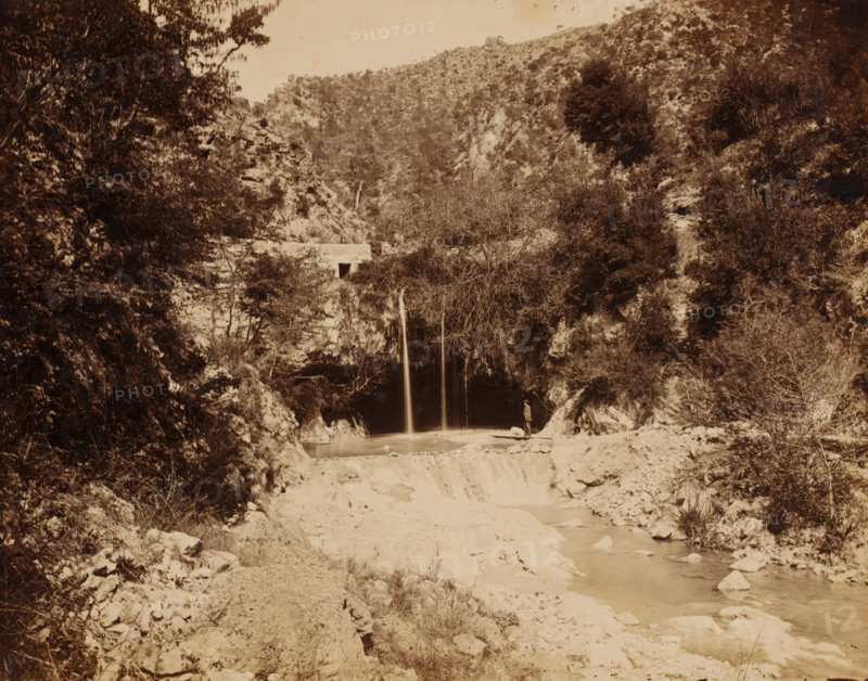Landscape with stream and waterfall, photograph, Landscape with stream and waterfall., c. 1880 - c. 1900, photographer, pasteboard, paper, albumen print, height 204 mm, width 270 mm x width 357 mm, height 298 mm, A sepia-toned landscape photograph showing a rocky riverbed and a narrow waterfall flowing into a pool surrounded by steep, vegetated canyon walls, with dense shrubs and trees on the slopes and a small bridge or concrete structure visible above the waterfall; a solitary figure stands on a rocky ledge near the pool, and the foreground shows a sandy, stone-strewn riverbank leading to the water.