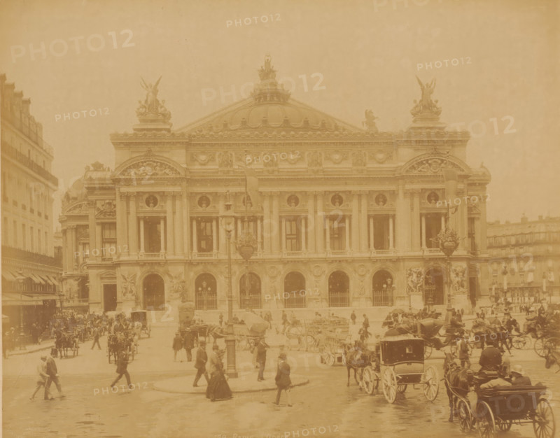 Opéra, Paris, photograph, Paris., inscription: '139 Paris, l'Opéra.', check stamp: 'Photographie Company, J. KUHN, Paris ... Rue de Rivoli ...', photographer: J. Kühn (publisher), c. 1885 - c. 1910, J. Kühn (publisher), photographer, paper, albumen print, height 220 mm, width 275 mm x width 397 mm, height 343 mm, Opéra de Paris, A large ornate stone building with columns and arched openings fills the center of the image, topped by a domed roof and sculpted figures on the roofline; flags hang from poles in front of the façade. In front of the building a busy street scene shows many horse-drawn carriages and people walking, some wearing long coats and hats and a few carrying umbrellas. Lamp posts and decorative sculptural elements are visible near the entrance, and adjacent buildings flank the main structure on either side. The whole scene appears in a single warm, sepia tone.