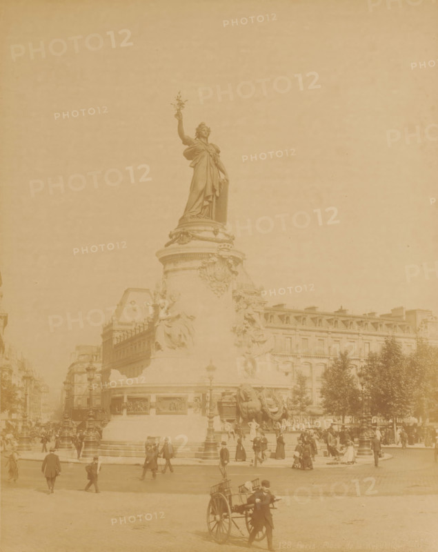 Place de la République, Paris, France, photograph, France., check stamp: 'Photography Company, J. KUHN, 220 170 174 Rue de Rivoli', inscription: '128 Paris. Place de la Republique', publisher: J. Kühn (publisher), c. 1885 - c. 1910, paper, albumen print, height 276 mm, width 218 mm x width 342 mm, height 402 mm, A large stone monument topped by a standing female statue holding a branch dominates the center, with sculpted reliefs and seated figures around its cylindrical base; surrounding the monument are multi-story buildings with rows of windows and mansard roofs, streetlamps, trees, and a crowd of people dressed in period clothing walking and gathering around the plaza, while in the foreground a man pulls a two-wheeled cart across the open paved square.