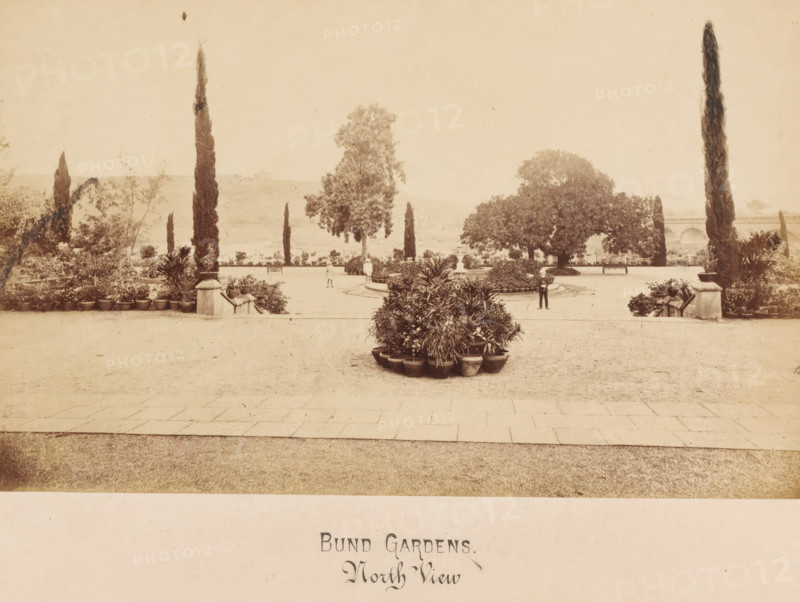 Bund Gardens, Pune, India, photograph, Oval photograph of a bridge with a river and a park on the left. Pune, also known as Pune and Puna and formerly Punavadi, is an Indian city not far from Bombay in the Western Ghats. The city is located in the district of Pune, which shares the same name., inscription: 'BUND GARDENS, POONA., North View.', inscription: 'Bund ..., North View', c. 1875 - c. 1900, photographer, paper, pasteboard, albumen print, height 146 mm, width 236 mm x width 358 mm, height 279 mm, A wide sepia-toned photograph of a formal garden with a paved foreground leading to a gravel area and a central display of potted plants arranged in a rounded cluster, flanked by raised stone planters and stairs on either side, tall narrow cypress trees rising at intervals, larger rounded trees and shrubs framing the middle distance, low hedges and additional potted plants along the terrace edge, a distant landscape with gentle hills visible beyond the garden, and a few small human figur...