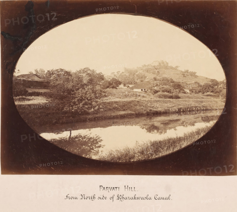 Parvati Hill from the north side of Rharakwasla Canal, India, photograph, India. The photo is elongated oval., inscription: 'Bund Gardens, South-West View', inscription: 'BUND GARDENS, POONA., South-West View.', c. 1875 - c. 1900, photographer, paper, pasteboard, albumen print, albumendruk, height 182 mm x width 246 mm, height 304 mm x width 393 mm, An oval-framed sepia landscape showing a hill with a cluster of trees and small buildings on its slopes, a body of water in the foreground reflecting the vegetation and hillside, grassy banks along the water, scattered trees and shrubs across the midground, and a dark vignette around the oval frame.