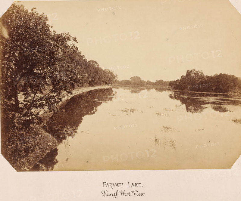 Parvati Lake, India. Viewed from the northwest, photograph, India. Viewed from the northwest., inscription: 'Parvati Lake, North-West', North-West View', c. 1875 - c. 1900, photographer, paper, pasteboard, albumen print, height 167 mm x width 239 mm, height 302 mm x width 393 mm, India, A sepia-toned photograph of a calm lake bordered by tree-lined banks, with dense foliage on the left foreground extending toward the middle distance and a narrower strip of trees on the right reflected in the still water, the shoreline curving gently and a low path or embankment visible along one side, scattered aquatic plants and reflections on the lake surface, and an expanse of pale sky above.