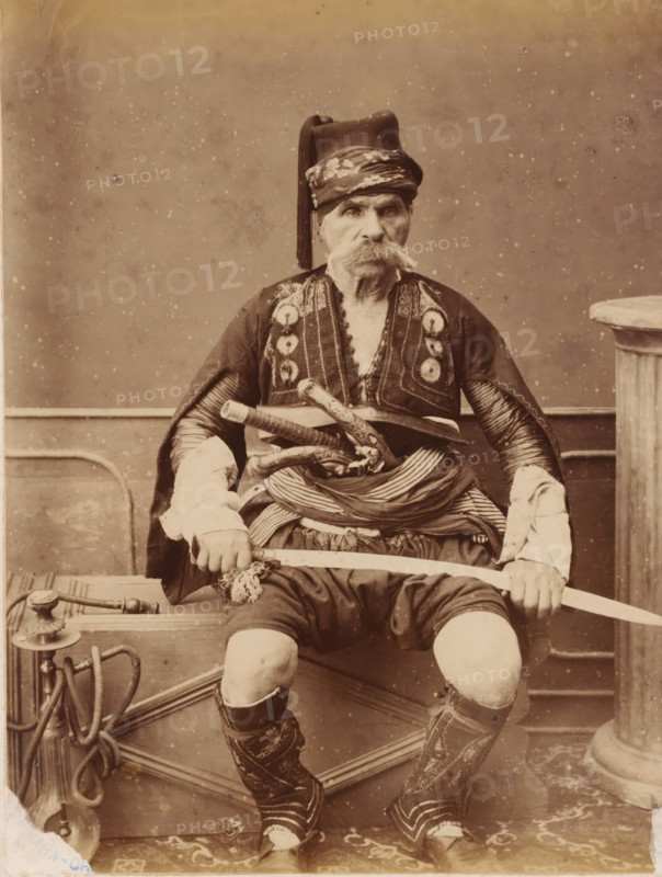 Portrait of old man in traditional costume, dervish?, photograph, dervish?., c. 1880 - c. 1900, photographer, paper, pasteboard, albumen print, height 228 mm, width 173 mm x width 389 mm, height 295 mm, An older man is seated facing forward, wearing a decorated dark jacket with patterned trim and a striped cloth wrapped around his waist, a tall hat with a long tassel, and high embroidered boots; he has a large bushy mustache and holds a curved sword horizontally across his lap with both hands, while several ornamental daggers or short blades are tucked into his waist sash and a coiled hose-like object and metal fixture sit beside the bench he occupies.