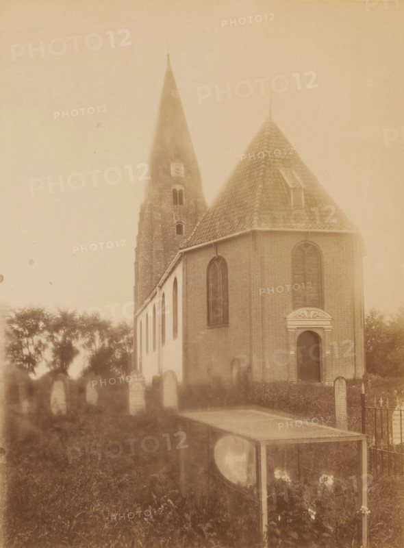 View of the St. Anthony's Church in Surhuizum, photograph, annotation: 'Surhuizen (Friesland), Dutch Reformed Church Sept. 1902, Achtkarspelen', annotation: 'Surhuizum, municipality of Achtkarspelen', photographer: AJM Mulder (possibly), Surhuizum, 1902, AJM Mulder, photographer, Attributed to, paper, pasteboard, albumen print, height 225 mm x width 169 mm, church (exterior), A sepia-toned photograph showing a brick church with a steep tiled roof and a tall pointed tower rising behind it, arched windows along the side of the building, an arched doorway with decorative stonework above it, rows of upright headstones in front of the church, a low metal fence section, trees in the background, and a rectangular glass-topped case or display box in the foreground standing on slender legs with flowers visible beneath it.
