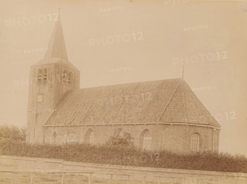 Former Reformed Church in Oosterwierum, photograph, annotation: 'Oosterwierum (Frieschland), NH Church Aug 1902, Baarderadeel', annotation: 'Oosterwierum, Oosterwierum, gem. Baarderadeel', photographer: AJM Mulder (possibly), 1902, Attributed to, paper, pasteboard, albumen print, albumendruk, height 166 mm x width 230 mm, church (exterior), A sepia-toned photograph showing a brick church with a steep tiled roof and a tall square bell tower topped by a pointed spire; the tower has arched openings and a circular feature on its face, and the nave displays arched windows along the side; a low hedge runs in front of the building and a stone wall with a railing is visible in the foreground.