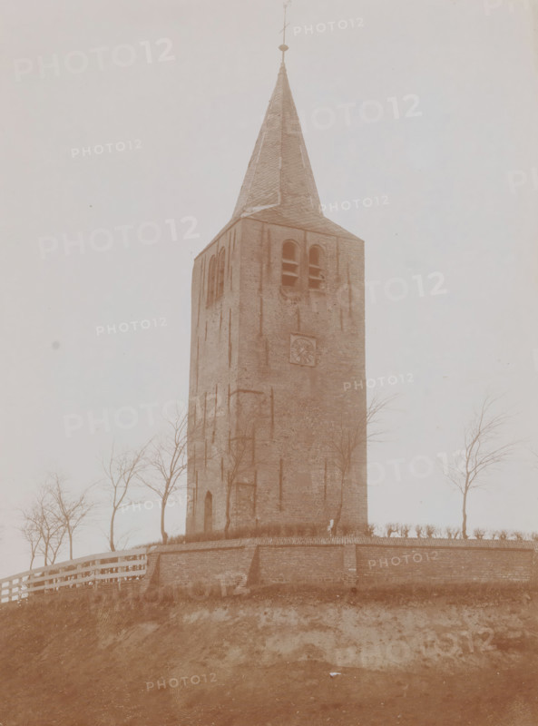 Tower of the Reformed Church in Oosterwierum, demolished in 1905, photograph, annotation: 'Oosterwierum, Oosterwierum, gem. Baarderadeel', annotation: 'Toren der formeren, NH Kerk te Oosterwierum, (Fr), Baarderadeel', photographer: anonymous (Monumentenzorg) (possibly), photographer: attributed to AJM Mulder, 1911, Attributed to, paper, pasteboard, daylight collodion silver print, height 219 mm x width 163 mm, parts of church exterior and annexes: tower, A tall brick tower with a steep, pointed roof topped by a metal finial rises from a low brick wall and grassy mound; the tower shows arched openings near the top with visible interior shelves or ledges and a circular carved stone feature on one face; bare trees stand around the base of the tower and a wooden fence runs along the top of the wall; the foreground shows an earthen slope leading up to the wall and a small piece of light-colored debris on the ground.