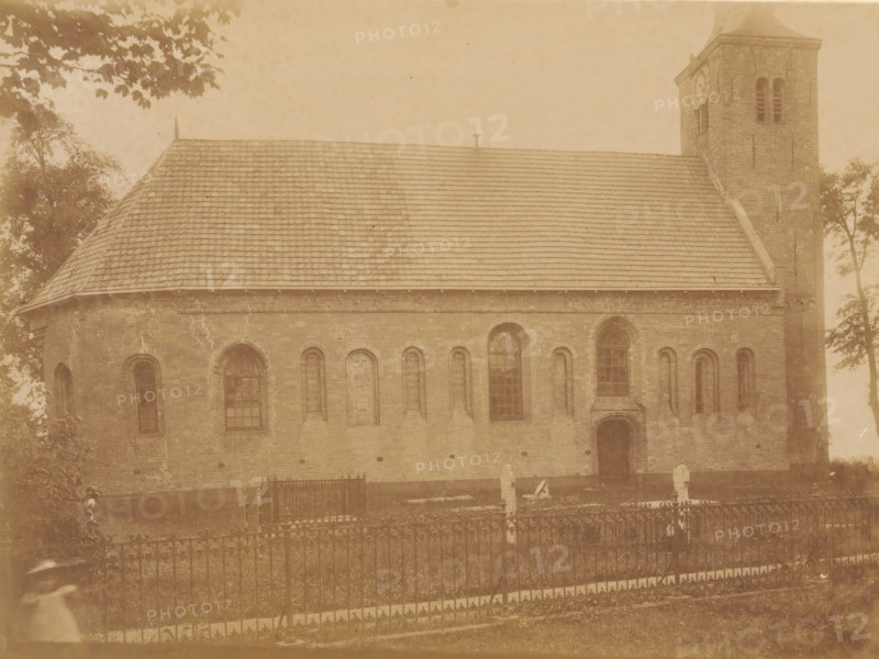 View of the Johanneskerk in Weidum, photograph, annotation: 'Weidum, Weidum, Baaderadeel', annotation: 'NH Church in Weidum (Friesland), list 1902, I. 278 No. 1394, Baarderadeel', photographer: AJM Mulder (possibly), 1902, Attributed to, paper, pasteboard, albumen print, albumendruk, height 168 mm x width 225 mm, church (exterior), Johanneskerk (Weidum), A large brick building with a steep tiled roof and a square tower is visible, showing a row of arched windows and a single wooden door set into the wall, with masonry details and weathering on the brick surface; a wrought iron fence runs across the foreground enclosing a grassy area with a few upright stone markers and a small fenced plot, while trees and overhanging branches frame the scene at the top and sides and a blurred figure appears near the lower corner of the image.