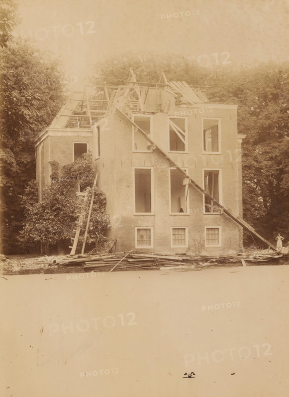 Dekema State in Weidum during the demolition, photograph, annotation: 'Weidum, Weidum, Dekema, gem. Baarderadeel', annotation: 'Weidum (Fr), Dekema State, list 1902', photographer: AJM Mulder (possibly), 1902, Attributed to, paper, pasteboard, albumen print, albumendruk, height 227 mm x width 166 mm, villa, A large brick building with its roof partially removed and wooden rafters exposed stands amid tall trees, the facade showing multiple window openings with some interior floors visible, slanted wooden beams propped against the front, scattered planks and debris piled at the base, a ladder leaning into foliage on one side, and a person standing near the building edge dressed in light clothing.