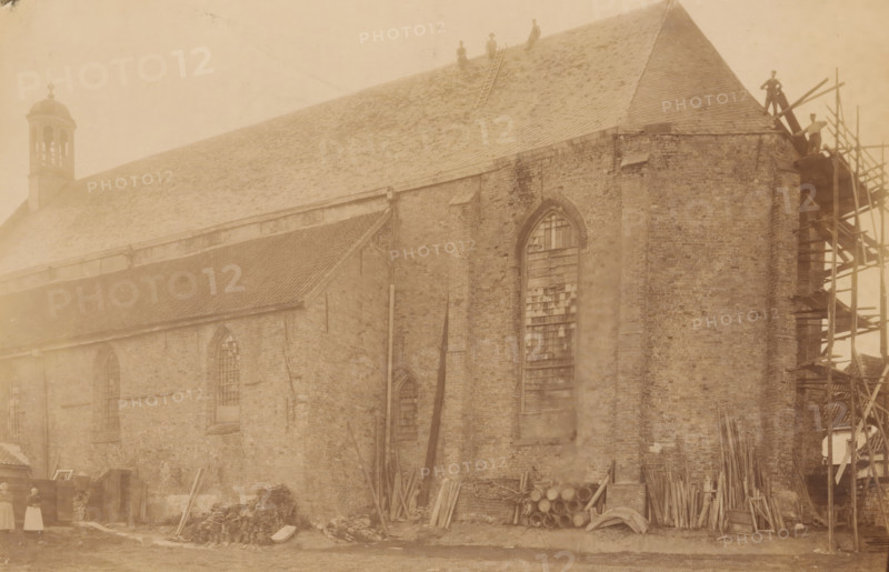 Broerekerk in Bolsward, during repair or restoration work, photograph, annotation: 'Bolsward (Fr), Broerenkerk, July 1903', annotation: 'Bolsward', Bolsward, photographer: AJM Mulder (possibly), 1903, Attributed to, paper, pasteboard, albumen print, height 151 mm x width 231 mm, church (exterior), A large brick building with a steep tiled roof fills the image, showing tall arched windows with broken or boarded panes, a small cupola near one end, and scaffolding against one corner where planks and beams are stacked; piles of wooden planks, beams, and stacked barrels lie along the base of the wall, and a ladder rests on the roof with several figures standing or sitting on the roof and near the scaffolding; the scene has a sepia tone and shows worn brickwork and scattered construction materials against a grassy foreground.