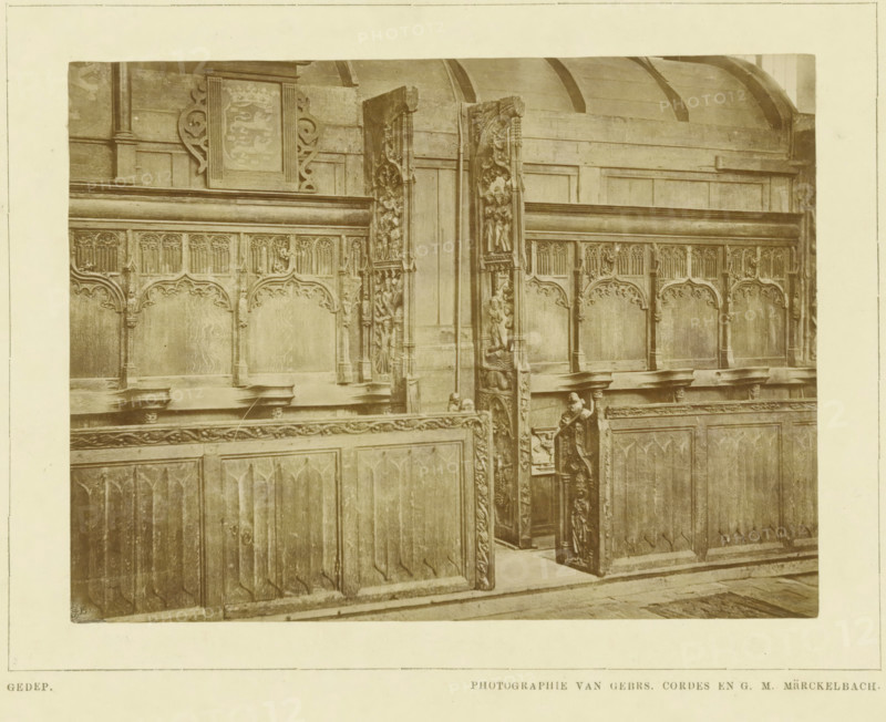 Choir stalls in the Martinikerk in Bolsward, photograph, inscription: 'Gedep., Photographie van gebrs. Cordes en GM Märckelbach', inscription: 'De Bolswardsche koorbanken, Ned. Kunstbode I, 1879 p.263', photographer: Cordes, Gebroeders, Bolsward, 1879, paper, pasteboard, albumen print, albumendruk, height 158 mm x width 229 mm, height 215 mm x width 280 mm, parts of church exterior and annexes: chapel, Martinikerk (Bolsward), The image shows a section of intricately carved wooden choir stalls with ornate panels and decorative arches along the upper portion, including carved vertical end pieces with figurative and floral reliefs; a narrow aisle between stalls is visible with a carved stall door partially open, revealing more carved details on its inner face; the wood has a worn, aged appearance with visible grain and patina, and a curved wooden ceiling element is visible above the stalls.