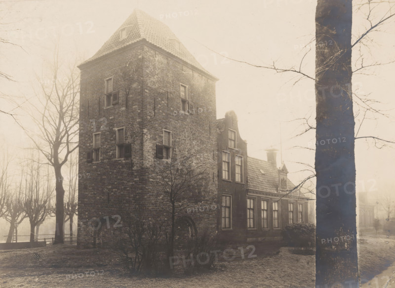 Residential tower Schierstins in Veenwouden, photograph, annotation: 'Dantumadeel, Veenwoude', annotation: 'Veenwouden (Fr), Schierstins restored, 21 Dec 1905', Veenwouden, photographer: AJM Mulder (possibly), 1905, Attributed to, paper, pasteboard, gelatin silver print, height 164 mm x width 224 mm, villa, A tall brick building with a steep tiled roof stands partially obscured by bare trees and a large tree trunk in the foreground; the building shows multiple small windows with shutters and an arched lower opening, and attached to it is a lower structure with larger rectangular windows and a decorative gable and chimney; leafless trees and low shrubs surround the structures and the scene is shrouded in a soft, pale light.