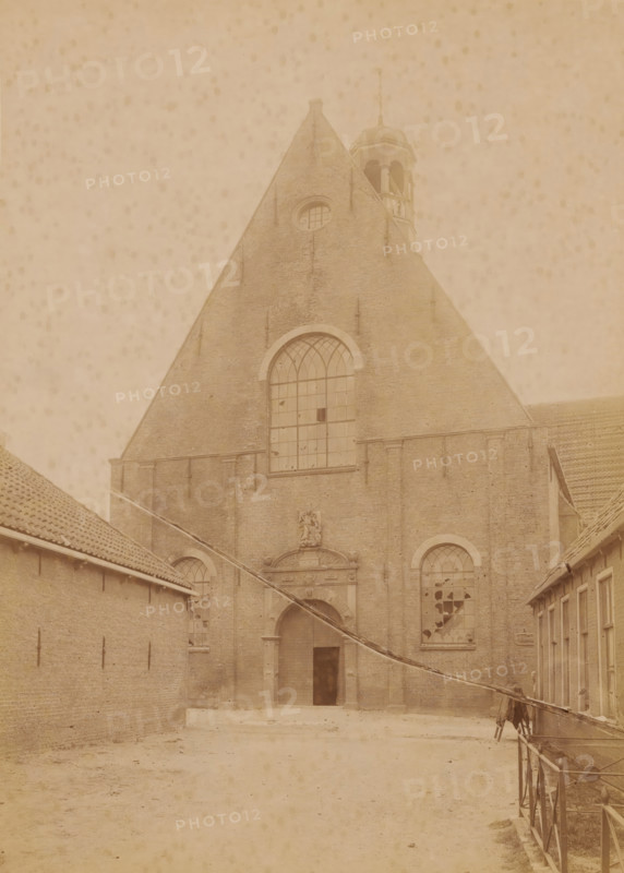 Westerkerk in Harlingen, shortly before its demolition in 1896, photograph, annotation: 'Harlingen', annotation: 'Westerkerk in Harlingen (Fr), July 1896', Harlingen, photographer: AJM Mulder (possibly), 1896, Attributed to, paper, pasteboard, albumen print, height 228 mm x width 165 mm, church (exterior), A large brick building with a steep triangular roof dominates the scene, featuring a tall arched window near the center and smaller arched windows lower on either side, some of which have broken panes; a decorative stone entrance with a carved crest sits beneath the central window and an open doorway leads into a dark interior; to the left and right, lower brick structures with tiled roofs flank a wide open courtyard that extends toward the foreground, where the ground appears bare and slightly uneven; a narrow railing runs along the right side of the courtyard and a slanted pole or beam crosses diagonally from the right toward the lower left; near the top of the main roof a small be...