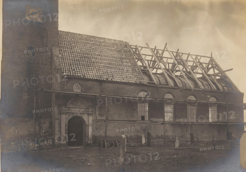 St. John the Baptist Church in Warns, during restoration work, photograph, annotation: 'Warns near Stavoren (Fr.), Dutch Reformed Church, Oct 1907', annotation: '..., Warns', Warns, photographer: AJM Mulder (possibly), 1907, Attributed to, paper, pasteboard, gelatin silver print, height 159 mm x width 225 mm, church (exterior), St. John the Baptist Church (Warns), A large brick building with a tall tower at one end occupies the scene, showing a steep tiled roof partially intact and partially removed to reveal a wooden roof framework and curved interior beams; the tower has a small arched window and a square clock face near its top. The facade features a central arched doorway with decorative stone surround above it and several tall arched windows along the wall, some with broken panes. Scaffolding and horizontal pipes run across the exterior, and wooden planks, construction debris, and upright posts are scattered on the ground in front of the building beneath a cloudy sky.