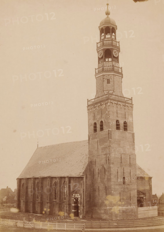 Northwest side of the Great Church in Hindeloopen, photograph, annotation: 'Hindeloopen, Church of the Dutch Reformed congregation, NW side, March 1892', annotation: 'Hindeloopen', Hindeloopen, photographer: AJM Mulder (possibly), 1892, Attributed to, paper, pasteboard, albumen print, height 238 mm x width 169 mm, church (exterior), A sepia-toned photograph showing a tall brick church tower rising from a large church building with a steeply pitched roof; the tower has multiple tiers with arched openings, decorative railings, a domed cupola topped with a finial, and visible clock faces, while the lower tower shows arched windows and buttresses; the side of the church displays tall lancet windows and an entrance with an ornamented surround; a wooden fence runs along the foreground and small adjacent buildings are partially visible at the edges.
