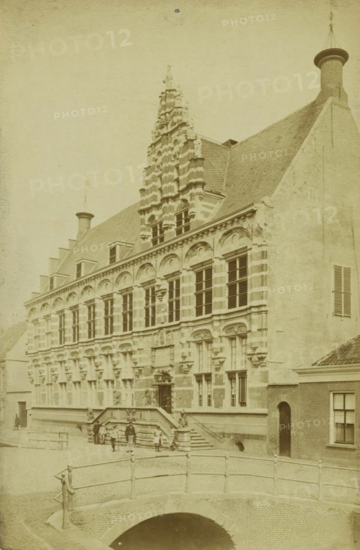 View of the chancery in Leeuwarden, photograph, inscription: '9 Leeuwarden sic. Nieuwe Stad', Leeuwarden, 1860 - 1900, photographer, paper, albumen print, height 182 mm x width 120 mm, façade (of house or building), A large historic stone building with ornate vertical facade details and rows of tall windows stands behind a short flight of steps leading to an entrance; the roof is steep and tiled with chimneys and decorative gables rising above, including a stepped central tower element with layered stonework, and adjacent plain wall sections with shuttered windows; in front of the building a stone balustraded bridge crosses a dark arched waterway, and several small figures and a few people stand or sit on the steps and near the bridge; the scene is rendered in a sepia-toned, aged photographic style.