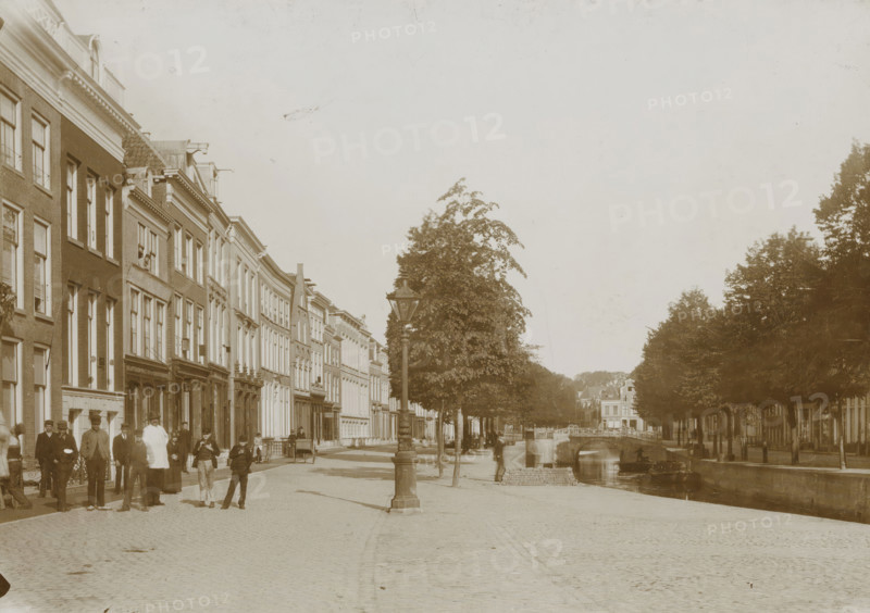 Street view in Leeuwarden, photograph, Leeuwarden, 1890 - 1920, photographer, paper, pasteboard, daylight collodion silver print, height 123 mm x width 174 mm, height 144 mm x width 198 mm, street (city(-scape) with figures, staffage), A wide cobblestone street runs alongside a canal with a stone bridge and row of trees, flanked on the left by a line of multi-story brick and stucco buildings with many tall windows and decorative facades, and on the right by dense tree foliage and more distant buildings; a tall ornate streetlamp stands near the canal edge, small boats sit on the water, and groups of people in period clothing stand and walk along the sidewalk near the buildings.