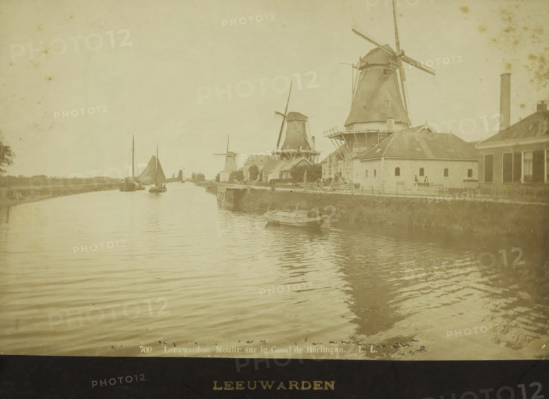 Mills along the canal from Leeuwarden to Harlingen, photograph, inscription: '700 Leeuwarden. Moulin sur le Canal de Harlingen. LL', inscription: 'LEEUWARDEN', photographer: Léon & Lévy, Friesland, 1864 - 1900, paper, pasteboard, albumen print, albumendruk, height 120 mm x width 183 mm, height 139 mm x width 200 mm, canal, windmill in landscape, A wide canal scene with calm rippled water in the foreground and a shoreline with grass and a low embankment on the right, several boats moored and sailing on the water including a small rowboat near the right bank and larger sailboats further back, a row of traditional windmills of varying sizes along the right-hand side behind low buildings and fences, pitched roofs and chimneys on the buildings, a tall smokestack near the right edge, a few figures standing near the buildings by the water, and an open sky above.