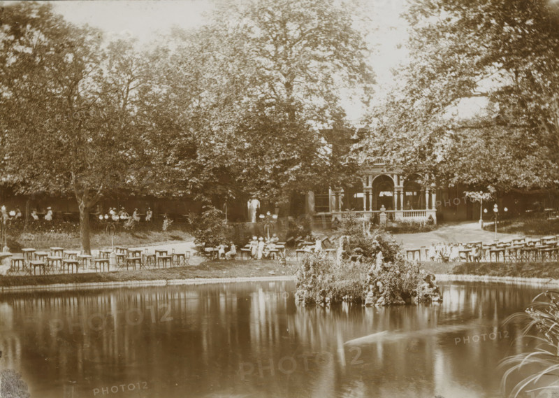 View of a park with a pond, presumably in Leeuwarden, photograph, Leeuwarden, 1890 - 1910, photographer, paper, pasteboard, daylight collodion silver print, height 124 mm x width 173 mm, height 144 mm x width 194 mm, park, A sepia-toned outdoor scene showing a pond in the foreground with a small rocky island covered in plants and flowers, its reflection visible in the water, and a grassy bank with scattered tables and chairs along the shore; beyond the seating area are large leafy trees partially obscuring a building with an ornate porch supported by columns, and people are seated at tables and standing near the structure, with lampposts and garden plantings interspersed among the trees.