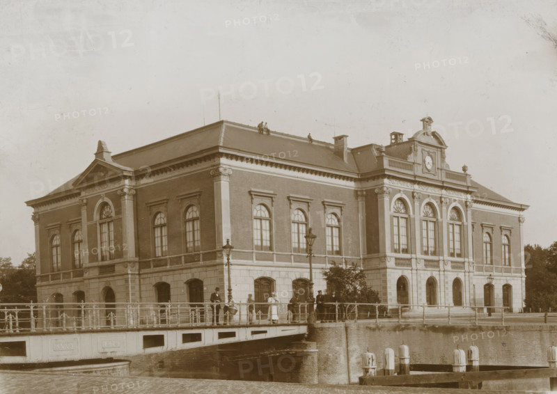 View of the former stock exchange building in Leeuwarden, photograph, Leeuwarden, 1890 - 1910, photographer, paper, pasteboard, daylight collodion silver print, height 122 mm x width 171 mm, height 144 mm x width 194 mm, Exchange, Bourse, A large stone and brick building with tall arched windows and decorative columns along its facade sits beside a canal, a sloped roof with small figures visible along the ridge, and a central pediment with a circular feature. A bridge with railings spans the water in front of the building, with lamp posts mounted along it and people standing and walking across, some wearing hats and long coats or dresses. The canal below the bridge has mooring posts and a stone retaining wall, and parts of the building's lower level show arched openings and a row of windows. The overall scene is in a monochrome sepia tone.
