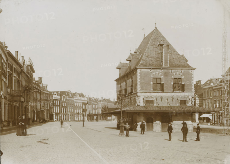 Street view with the Waag in Leeuwarden, photograph, photographer: anonymous, Leeuwarden, 1890 - 1910, paper, pasteboard, daylight collodion silver print, height 126 mm x width 174 mm, height 144 mm x width 199 mm, street (city(-scape) with figures, staffage), weigh-house, A wide cobblestone street lined with multi-story brick buildings leads to a large steep-roofed brick building with dormer windows and a covered arcade at its base; people stand and walk in the open area in front of the building, including men in dark suits and hats and a woman in a long dark dress with a hat, while lamp posts and a few small street-level awnings are visible along the street and façades of adjacent buildings.