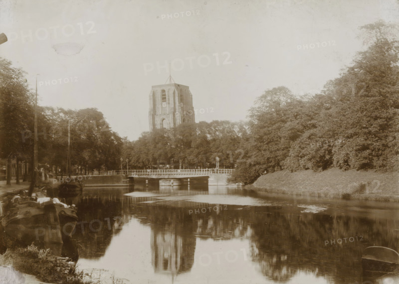 View of the Oldehove with canal in Leeuwarden, photograph, Leeuwarden, 1890 - 1910, photographer, paper, pasteboard, daylight collodion silver print, height 125 mm x width 173 mm, height 144 mm x width 194 mm, canals, waters (in city), parts of church exterior and annexes: tower, A sepia-toned riverside scene showing a tall stone tower rising above a row of trees, a low bridge crossing the river in front of the trees, reflections of the tower and trees visible on the water surface, boats moored along the near bank with some rigging and equipment, dense foliage on both sides of the river, a lamp post near the bridge, and a calm sky overhead.