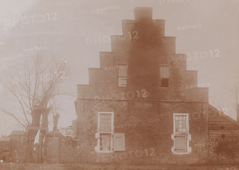 Aytta State (godshouse or church) in Swichum, photograph, annotation: 'Aytta Godshuis Swichem, Friesland, Dec. 1909, Leeuwarderadeel', Swichum, photographer: AJM Mulder (possibly), 1909, Attributed to, paper, pasteboard, daylight collodion silver print, height 151 mm x width 209 mm, church (exterior), A sepia-toned photograph of a stepped brick building with a central raised section and tiered sides, showing multiple windows with shutters and panes in varying states, two arched brick window surrounds on the lower level, and a doorway partially visible. Bare tree branches flank the building on both sides, and a wooden fence and gate are visible at the left base of the building. The brickwork shows weathering and variations in tone, and the overall scene has a faded, aged appearance.