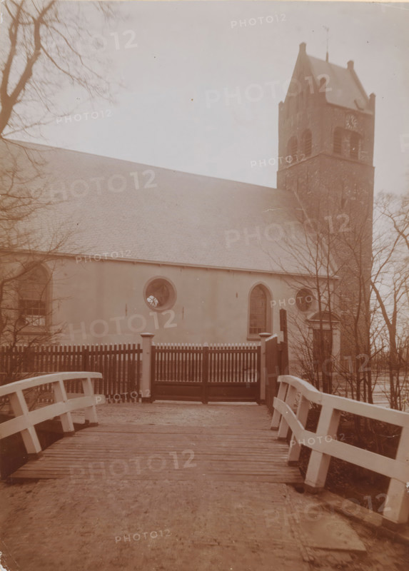 St. John the Baptist Church in Huizum, photograph, annotation: 'Dutch Reformed Church with tower in Huizum (Fr) Jan 1911, Leeuwarderadeel', Huizum, photographer: AJM Mulder (possibly), 1911, Attributed to, paper, pasteboard, daylight collodion silver print, height 216 mm x width 151 mm, church (exterior), A wooden bridge with painted railings leads to a gate with vertical slats and posts, beyond which stands a building with a long sloped roof and smooth walls. The building facade shows rounded and arched windows, one circular window with visible radial mullions and a taller arched window nearby. To the right a tall brick tower with arched openings and a steep roof rises above the building. Bare tree branches frame the scene on both sides and a pale sky fills the upper background.