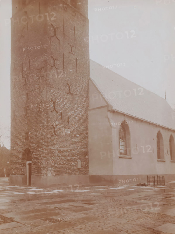 St. John the Baptist Church in Huizum, photograph, annotation: 'Detail of tower and Church of the Dutch Reformed Congregation in Huizum (Fr) Jan 1911, Leeuwarderadeel', Huizum, photographer: AJM Mulder (possibly), 1911, Attributed to, paper, pasteboard, daylight collodion silver print, height 218 mm x width 163 mm, church (exterior), A tall brick tower with visible metal anchors and an arched doorway sits adjacent to a plastered building with arched windows and a sloped roof, the ground in front paved with large rectangular stones and wet patches, and a small metal fence near the building's corner.