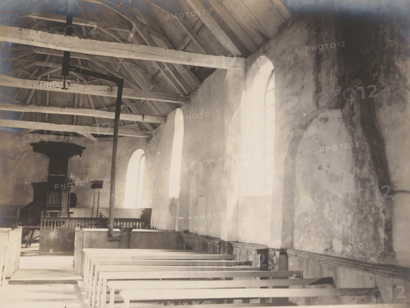 Interior of the Reformed Church in Siegerswoude, demolished in 1909, photograph, annotation: 'Siggerswoude friesland, Kerk der Ned: Herv: municipality, Oct 1908, Opsterland', Siegerswoude, photographer: AJM Mulder (possibly), 1908, Attributed to, paper, pasteboard, gelatin silver print, developing gelatin silver print, height 162 mm x width 216 mm, interior of church, An interior view showing wooden rafters and beams supporting a pitched roof, a row of tall arched windows letting bright light into a long nave lined with wooden pews, plastered walls with visible patches and discoloration, a vertical metal pipe running from floor to ceiling, a raised platform area with a dark wooden pulpit and railing, and various wooden paneling and architectural details along the lower walls.