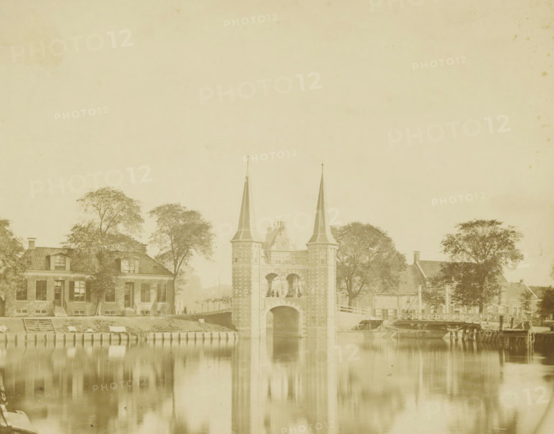 View of the Sneeker Waterpoort, photograph, annotation: '... f1, -, Sneek', photographer: anonymous, Sneek, 1870 - 1900, paper, pasteboard, albumen print, albumendruk, height 145 mm x width 182 mm, height 252 mm x width 283 mm, city-gate (city(-scape) with figures, staffage), Waterpoort (Sneek), A pale photograph shows a river with calm reflective water in the foreground and a brick gateway structure with two tall pointed spires rising above an arched passage at the center; flanking the gateway are trees and houses with pitched roofs and dormer windows, and a small bridge and wooden piers extend along the riverbank to the right, while the scene is framed by more trees and building facades on either side, all rendered in muted sepia tones.