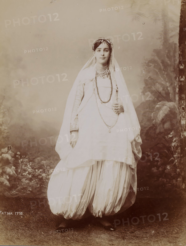 Portrait of a standing woman in front of a painted background, photograph, Portrait of a standing woman in a light robe in front of a painted background., inscription: 'FATMA 1773', c. 1880 - c. 1900, photographer, paper, daylight gelatin silver print, height 251 mm x width 190 mm, A full-length portrait of a young woman standing and facing forward wearing a flowing light-colored dress with a draped outer garment and a veil over her head, her dark hair visible at the forehead and adorned with a decorative headpiece, layered necklaces and long bead strands over her chest, long sleeves with visible cuffs, and a hand holding part of the drape near her waist; the skirt is voluminous and gathered at the bottom, revealing patterned stockings and dark pointed shoes at the feet; the background shows a studio backdrop with foliage motifs and a textured floor beneath her.