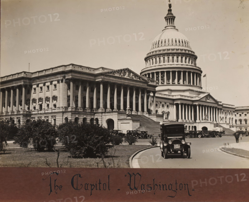 The Capitol, Washington DC, United States, album page, photograph, United States., inscription: 'The Capitol - Washington', c. 1900 - c. 1940, photographer, pasteboard, paper, albumen print, height 146 mm x width 218 mm, height 207 mm x width 263 mm, North America, District of Columbia, Capitol (Washington DC), A large neoclassical building with a prominent domed rotunda and a colonnaded façade fills most of the image, showing a wide staircase leading up to the entrance and sculptural details on the pediments, with rows of tall columns and ornate window treatments along the length of the structure; several early model automobiles are parked and driving on a curved roadway in front of the building, a taller vehicle is closest to the camera, and small trees and shrubs occupy the grassy area between the road and the building.