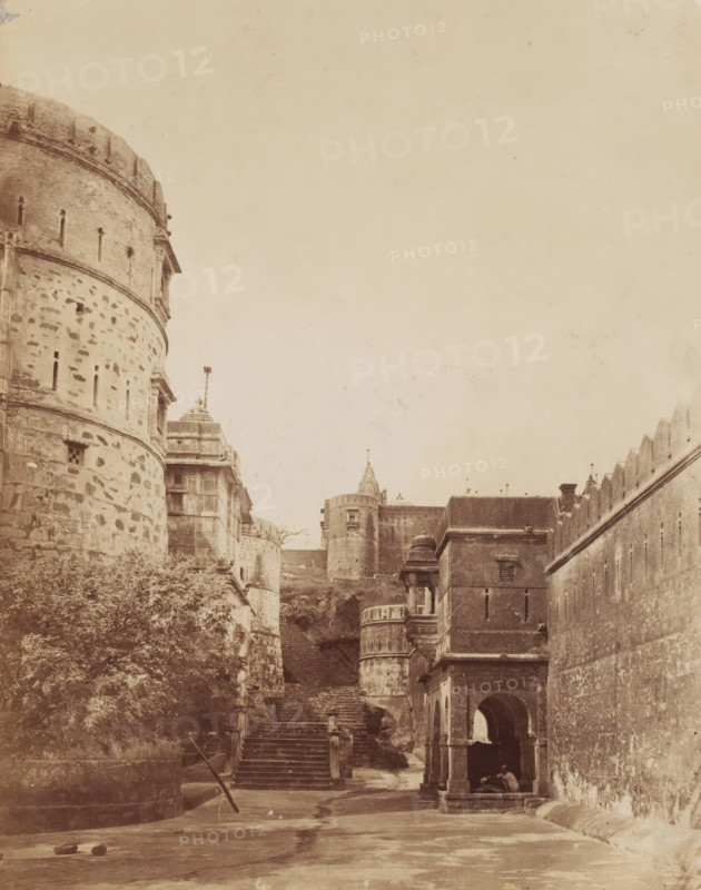 Temple complex at Palitana, Gujarat, India, photograph, Castle complex ?, India ?., Palitana, c. 1870 - c. 1900, photographer, paper, albumen print, height 232 mm x width 183 mm, A sepia-toned view of a stone fortress complex with rounded towers and high walls; a large cylindrical tower with small vertical slits and textured stonework occupies the left foreground, while a series of steps lead uphill between buildings toward additional towers and battlements in the midground; on the right a tall wall with crenellations runs diagonally, and nearer to the viewer a square stone pavilion with arched openings and a domed roof sits beside the steps; vegetation grows at the base of the left tower and two small objects lie on the ground in the open courtyard area in the foreground.